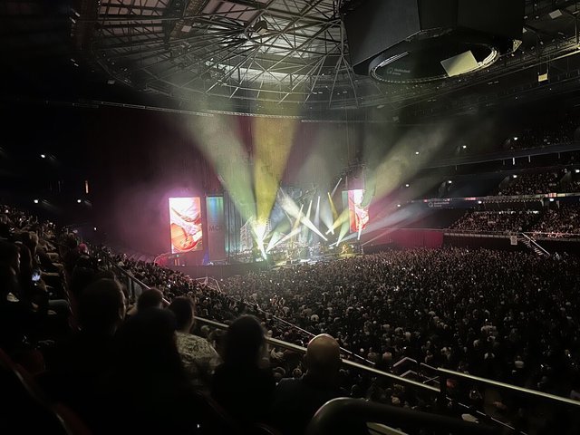 Wide view of an arena concert with colored stage beams, video screens and a packed floor