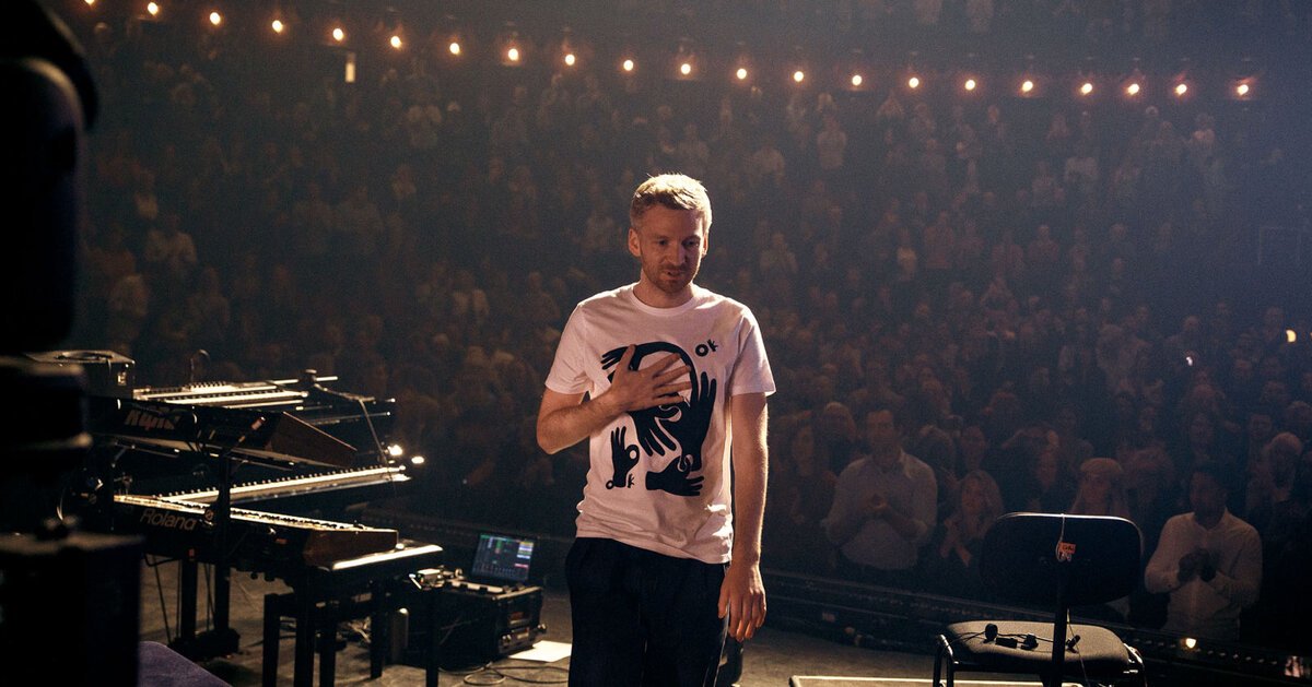 Ólafur Arnalds standing on stage beside keyboards and a piano, facing a packed, applauding theater