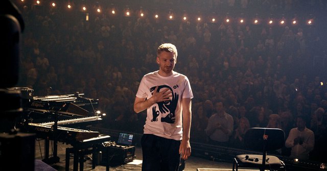 Ólafur Arnalds standing on stage beside keyboards and a piano, facing a packed, applauding theater