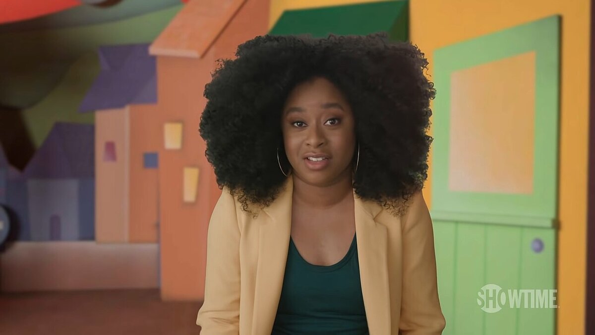 Phoebe Robinson in a yellow blazer and green top, smiling slightly in front of a colorful set