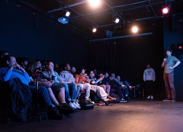 Audience seated in a dark black-box theater watching a speaker with a microphone under stage lights