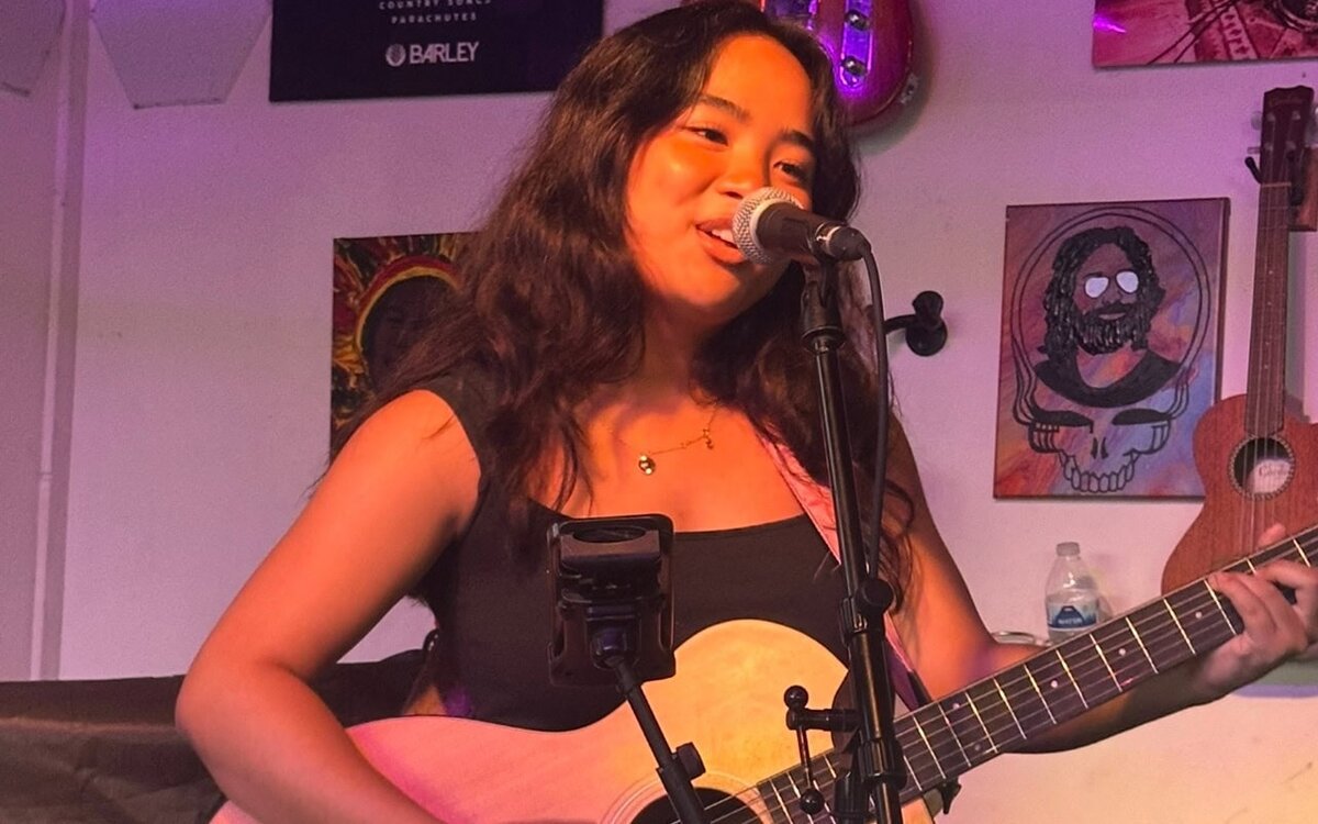 Singer-songwriter with an acoustic guitar performing at a microphone on a small stage with colorful posters and a ukulele on the wall behind her