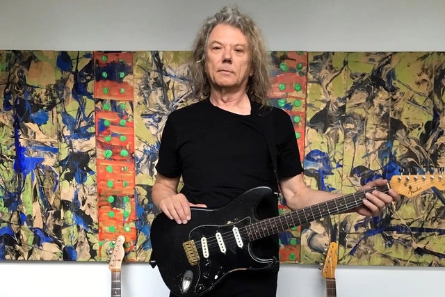 Jerry Harrison holding a black electric guitar in front of an abstract painting