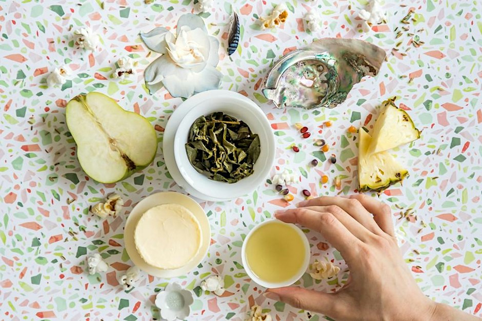 A white gaiwan of steeped oolong leaves and a small cup of pale tea on a terrazzo cloth, surrounded by a halved pear, pineapple wedges, a gardenia and an abalone shell