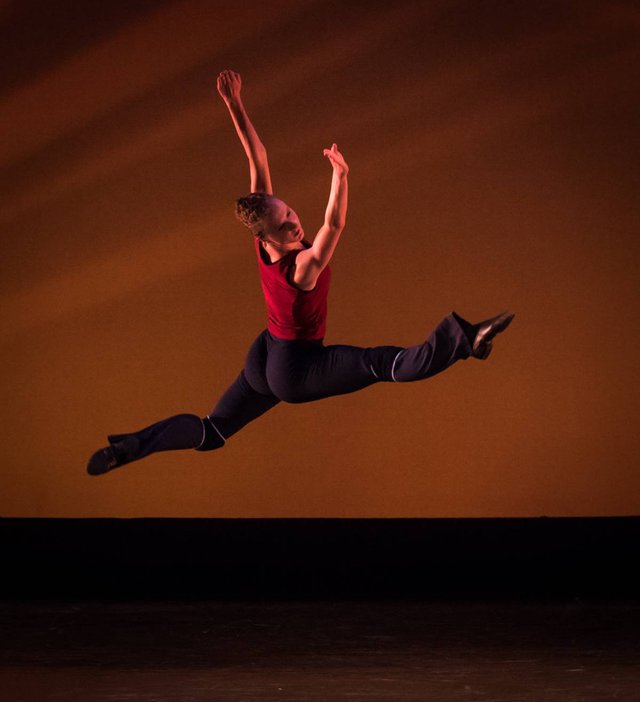 Dancer in a red top leaping mid-air against an amber stage backdrop