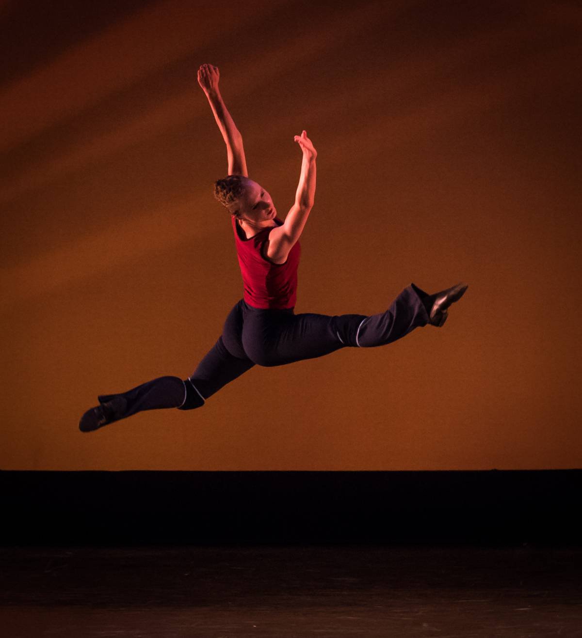 Dancer in a red top leaping mid-air against an amber stage backdrop