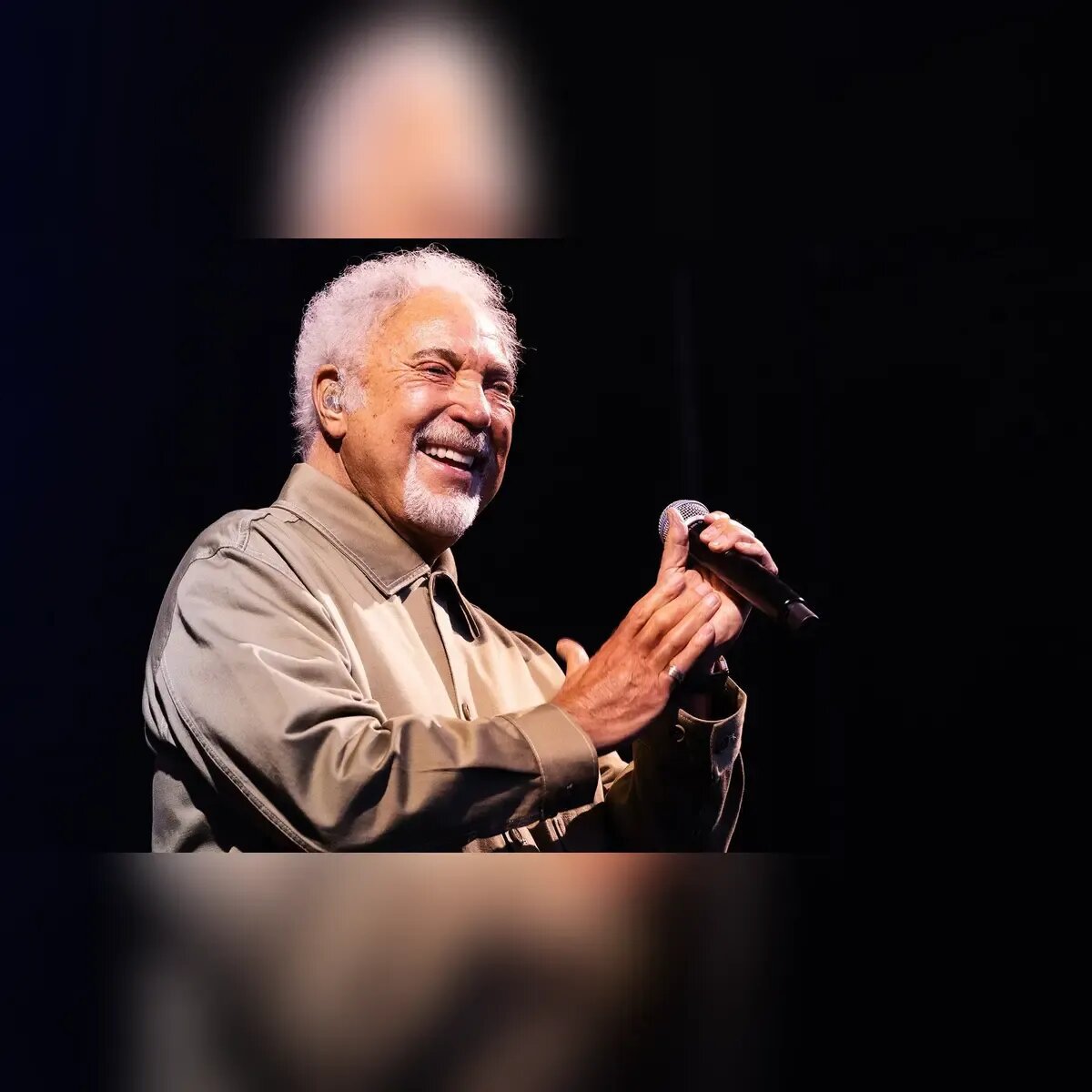 Tom Jones smiling on stage with a microphone in both hands against a dark backdrop