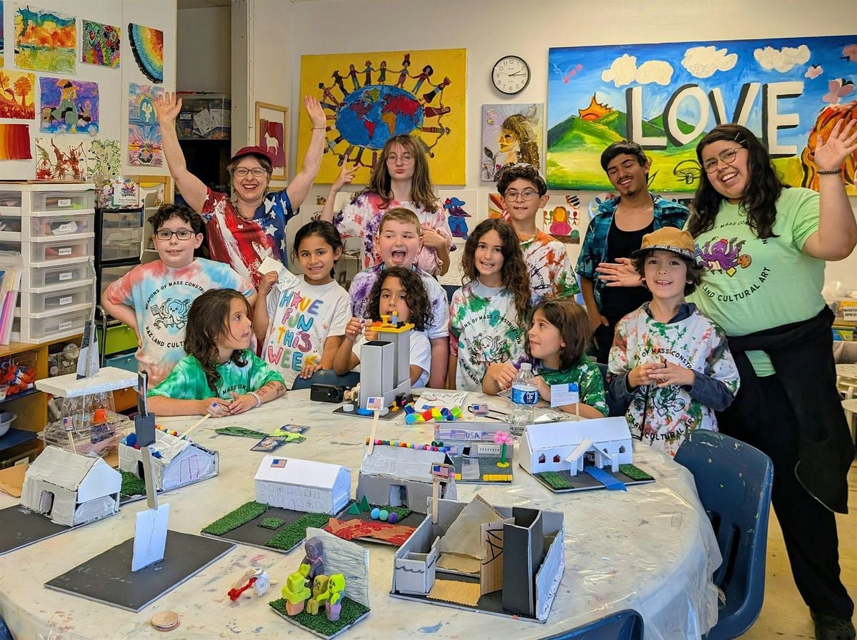 Smiling kids and two adult leaders in tie-dye shirts around a worktable of small cardboard model buildings in an art studio