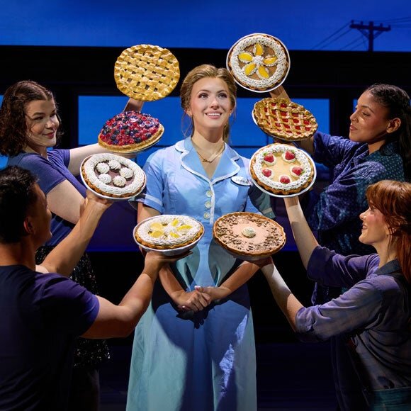 A smiling waitress in a light blue uniform surrounded by cast members holding up decorated pies on a stage