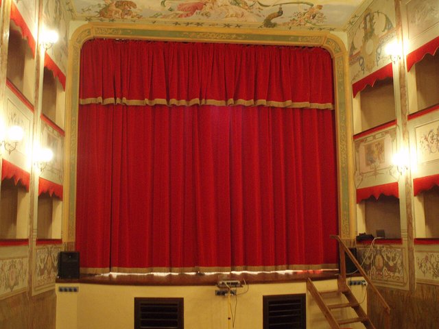 An empty small theatre stage behind a red curtain, framed by tiers of boxes