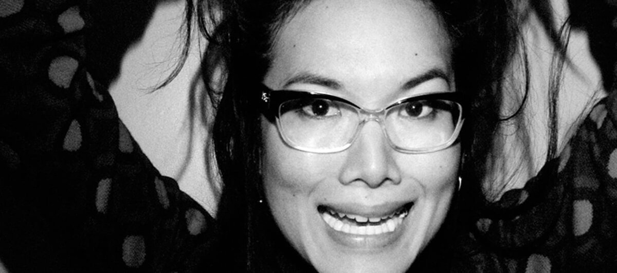 Black and white close-up portrait of a smiling woman in clear-framed glasses with her arms raised