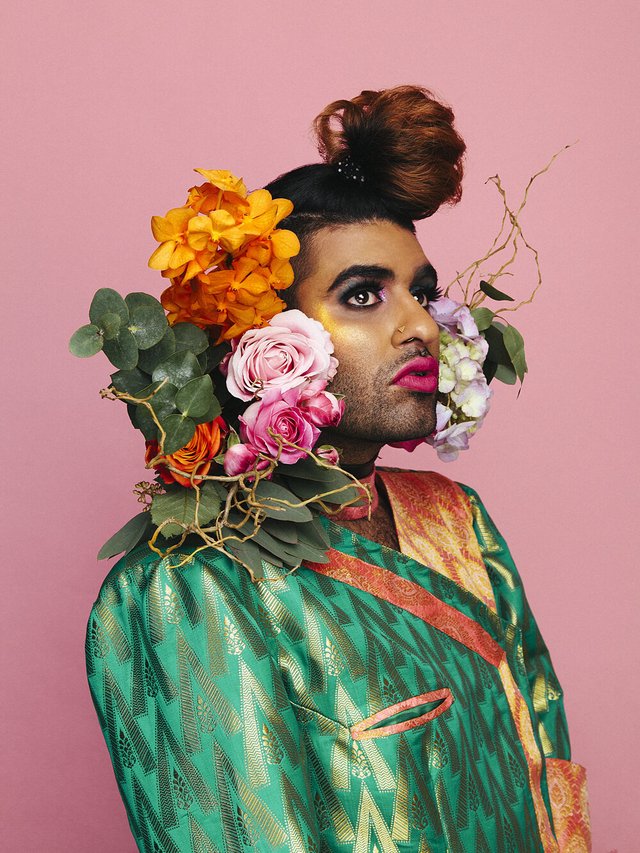 Portrait of Alok Vaid-Menon against a pink backdrop, wearing a green brocade jacket with flowers at the shoulder