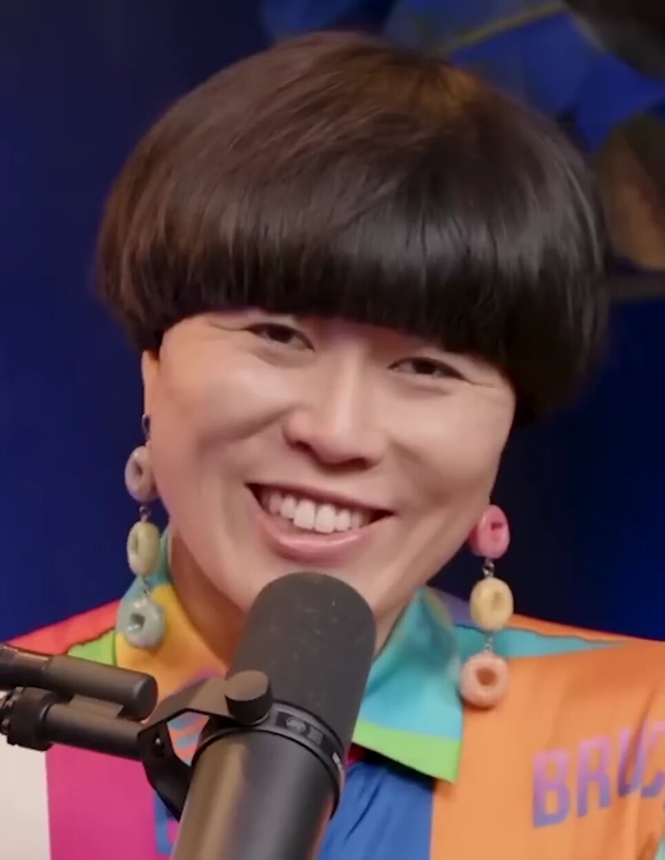 Atsuko Okatsuka smiling at a microphone in a colorful jacket and cereal-ring earrings
