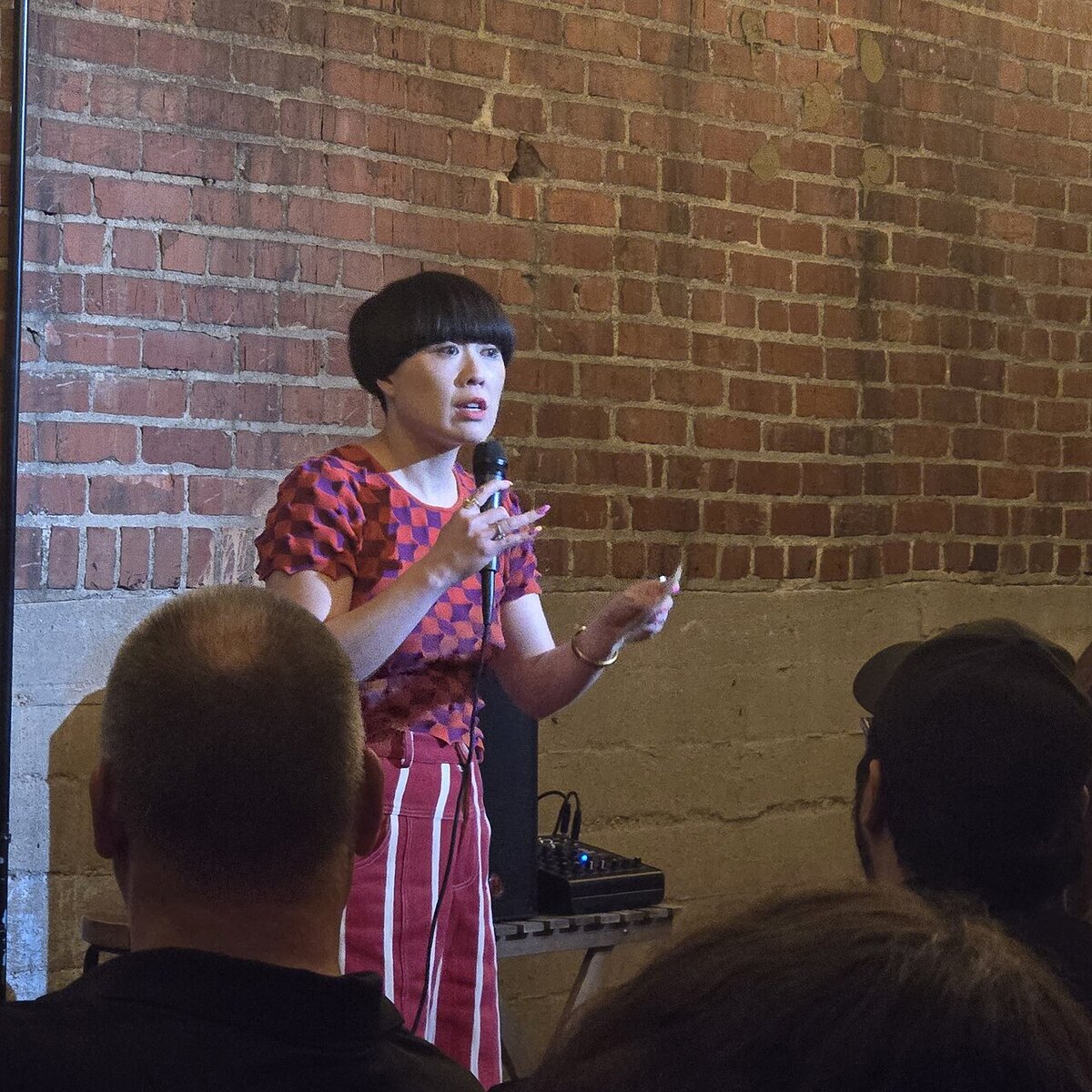 Comedian Atsuko Okatsuka holding a microphone on stage in front of a brick wall, with audience heads in the foreground