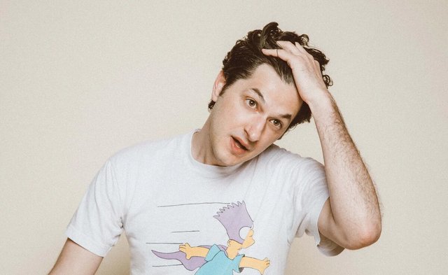 Portrait of a dark-haired man in a white graphic t-shirt running a hand through his hair against a cream backdrop