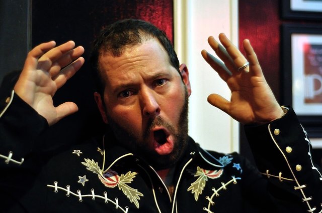 Bert Kreischer in an embroidered black western shirt, hands raised and mouth open mid-story
