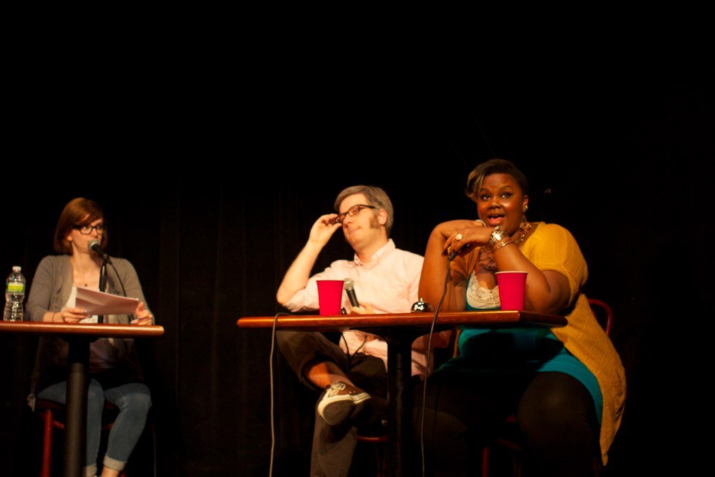Nicole Byer seated at a table on a dark stage during a live comedy show