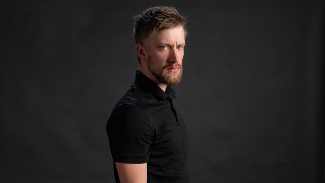 Daniel Sloss in a black polo shirt, glancing sideways at the camera against a dark studio backdrop