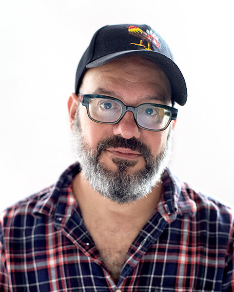 Portrait of a bearded man in glasses, a dark cap and a plaid shirt against a white background