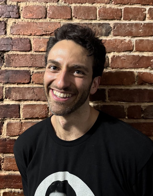 Gianmarco Soresi smiling in a black t-shirt in front of a red brick wall