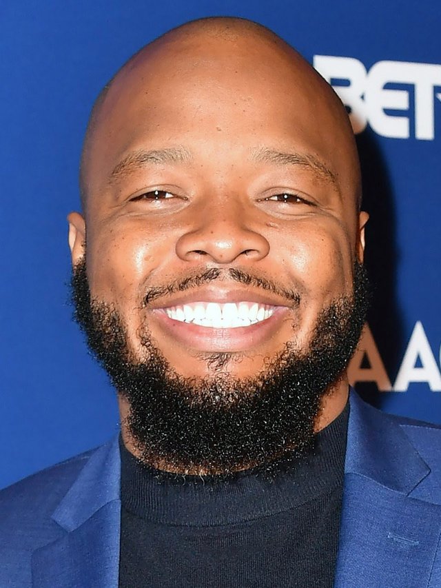 KevOnStage smiling in a blue blazer and black turtleneck in front of a blue step-and-repeat backdrop