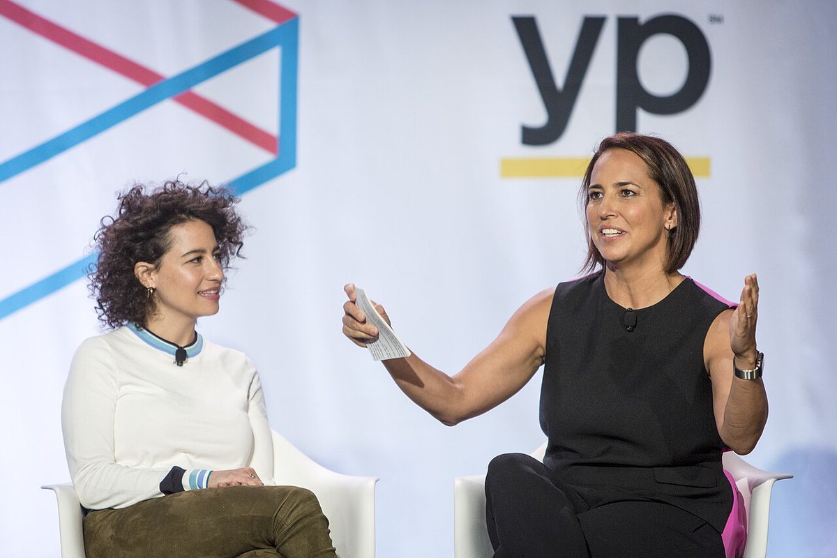 Ilana Glazer, with curly dark hair and a white sweater, seated onstage during a panel conversation
