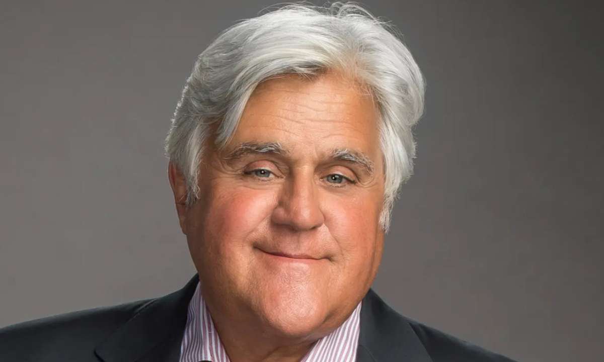 Portrait of Jay Leno with silver hair, smiling, in a dark jacket against a gray backdrop