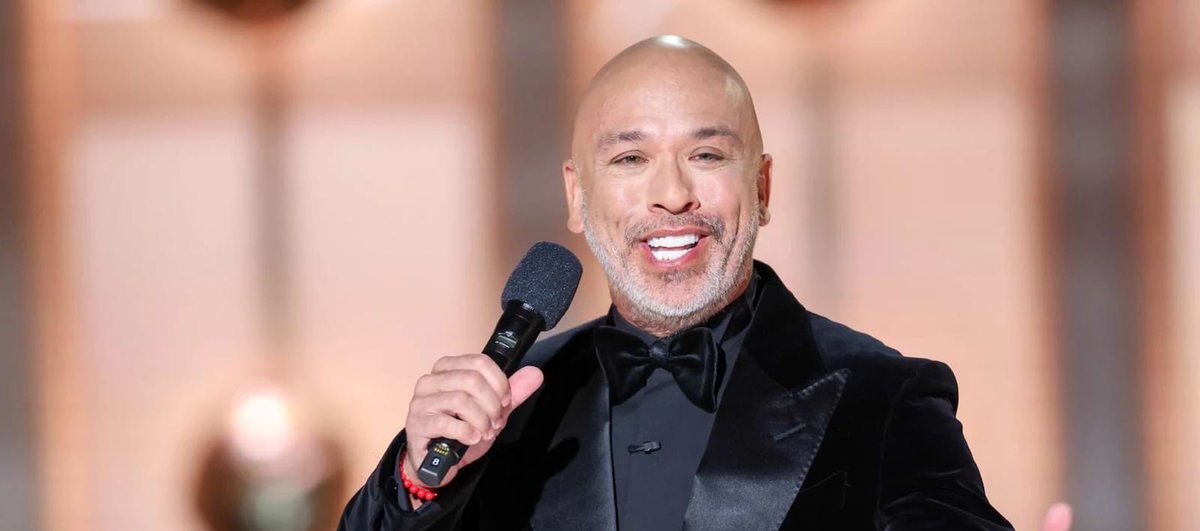 Jo Koy smiling and holding a microphone while wearing a black velvet tuxedo jacket and bow tie