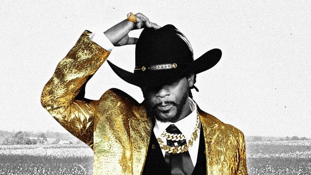 Katt Williams in a gold metallic jacket and black cowboy hat, tipping the hat over his eyes