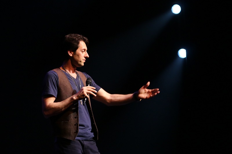 Max Amini on a dark stage holding a microphone and gesturing toward the audience under two spotlights