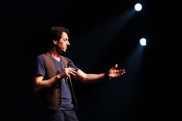 Max Amini on a dark stage holding a microphone and gesturing toward the audience under two spotlights