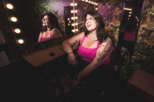 Cara Meyers in a pink tank top smiling in a dressing room with lit vanity mirrors