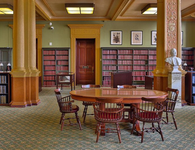 A quiet library reading room with a large wooden table and chairs, bookshelves along the walls and tall windows