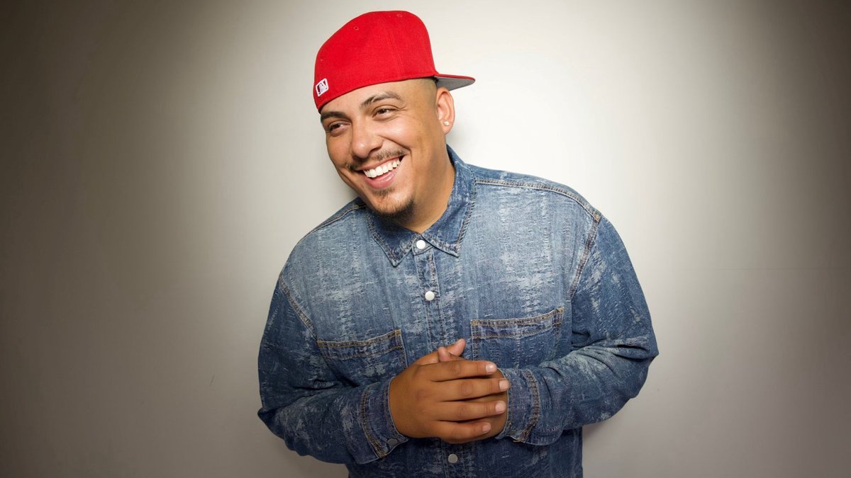 Concrete smiling in a red baseball cap and a denim button-up shirt against a light gray wall