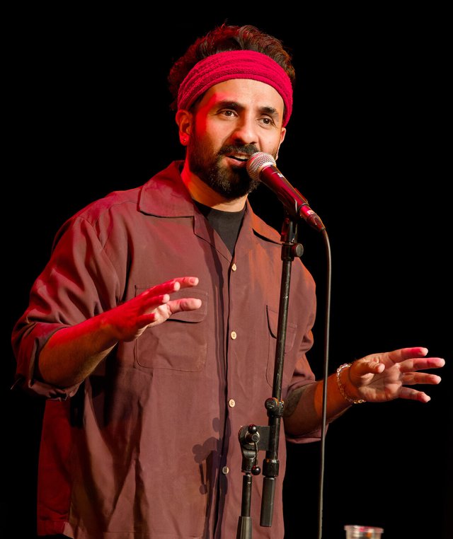 Vir Das in a red headband and maroon shirt speaking into a microphone on stage