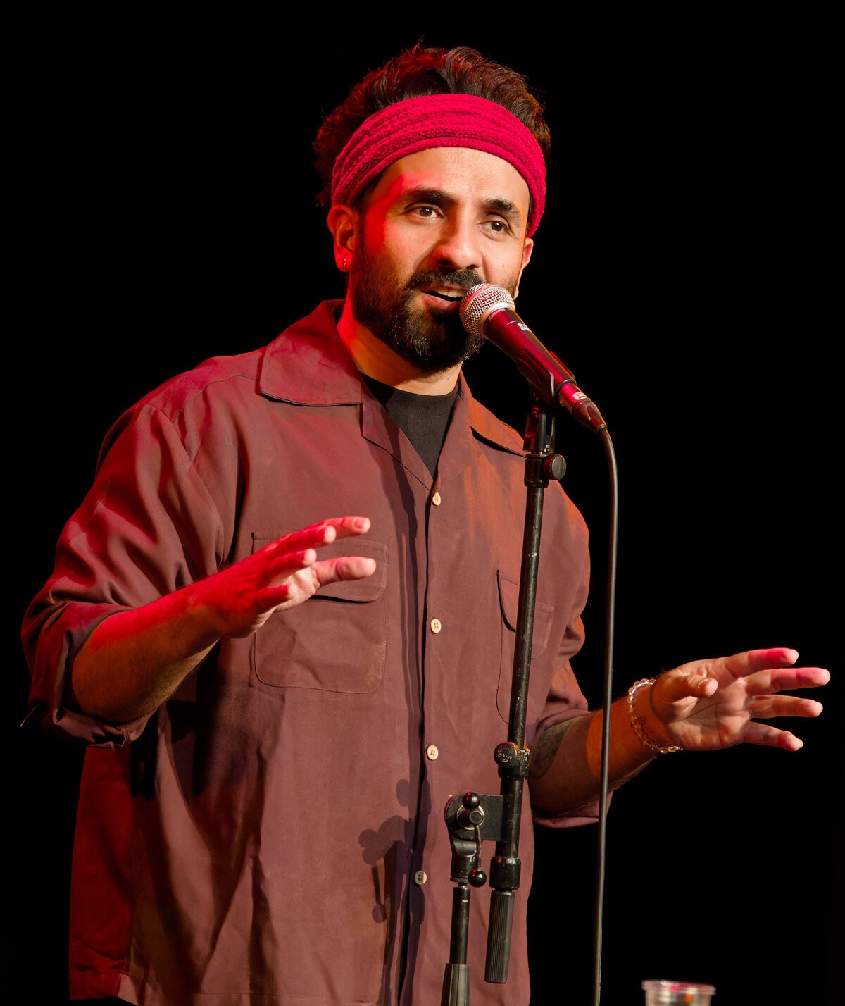 Vir Das in a red headband and maroon shirt speaking into a microphone on stage