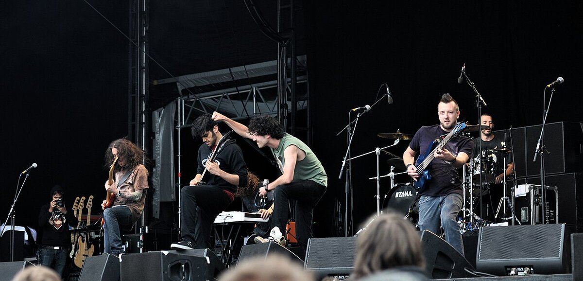 Metal band performing on an outdoor festival stage, guitarists and a bassist in front of a drum kit