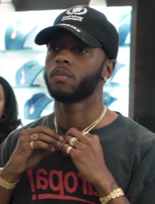 Portrait of 6LACK in a black cap and gold chains, adjusting his necklace