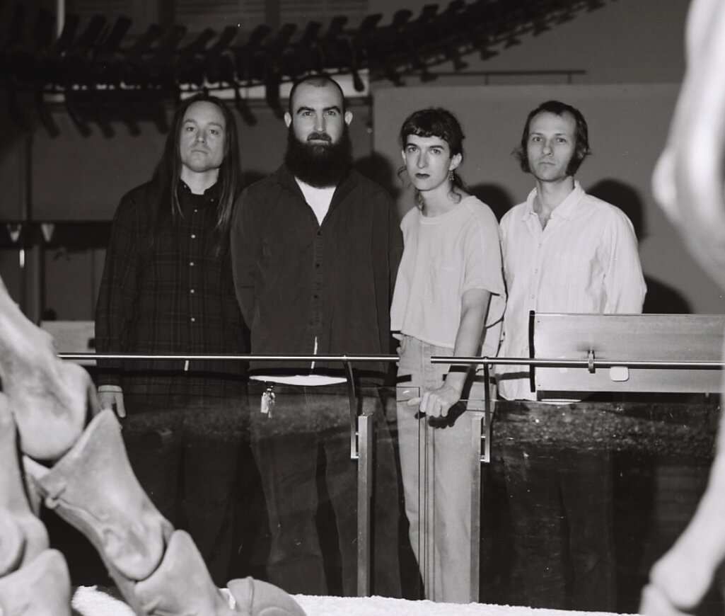 Black-and-white photo of four band members standing behind a glass railing in a museum, with large fossil bones in the foreground