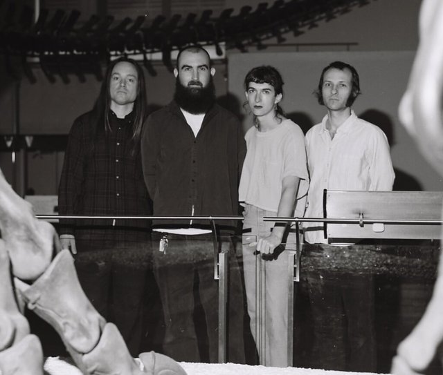 Black-and-white photo of four band members standing behind a glass railing in a museum, with large fossil bones in the foreground