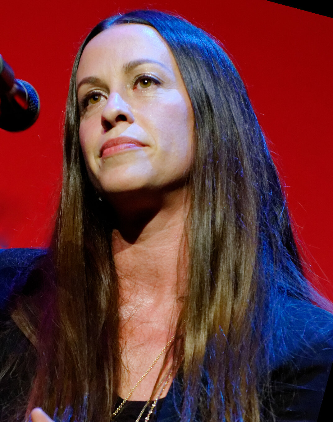 Close-up of Alanis Morissette singing at a microphone against a red stage backdrop