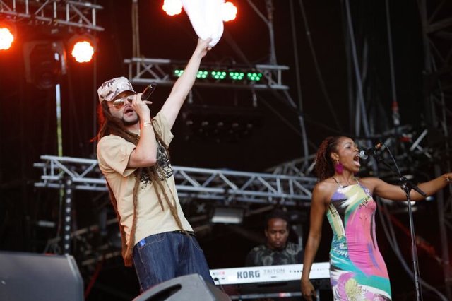 Alborosie in a cap and sunglasses raising a microphone on stage, with a backing vocalist and keyboardist beside him