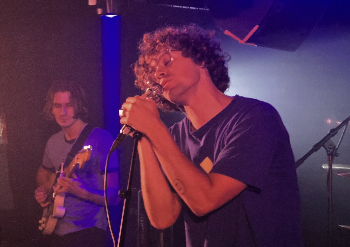 Almost Monday's singer holding the microphone close with a guitarist behind him under purple stage light
