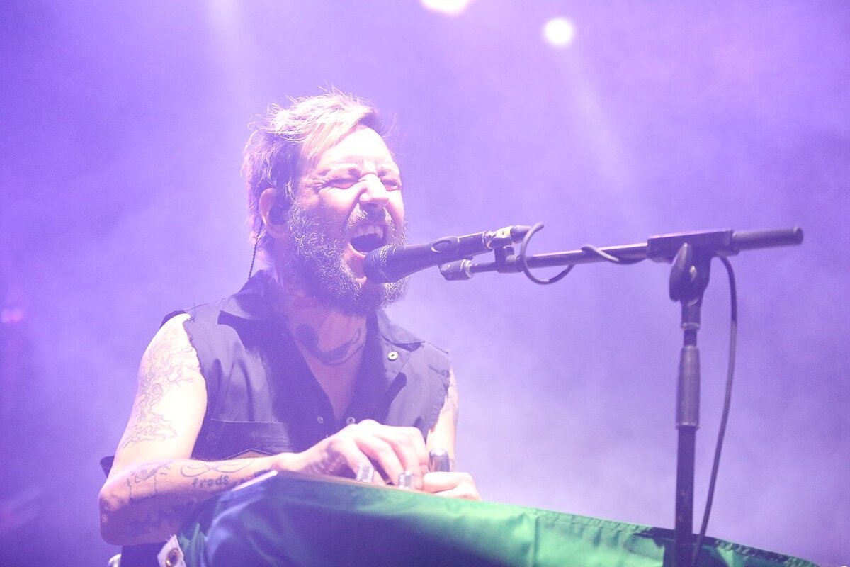 Bearded singer mid-song at a microphone, lit in purple stage haze