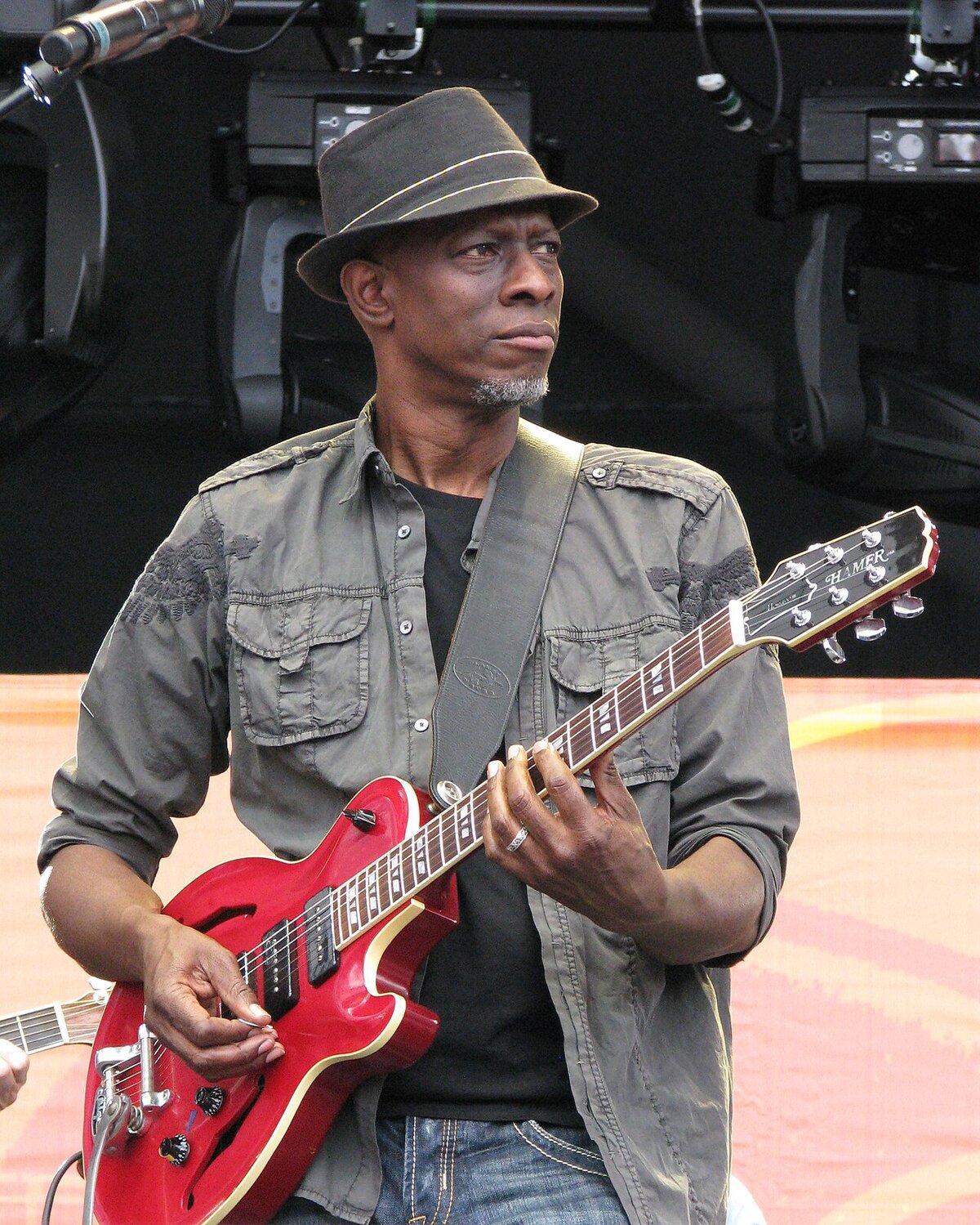 Keb' Mo' in a brimmed hat playing a red semi-hollow guitar on an outdoor stage