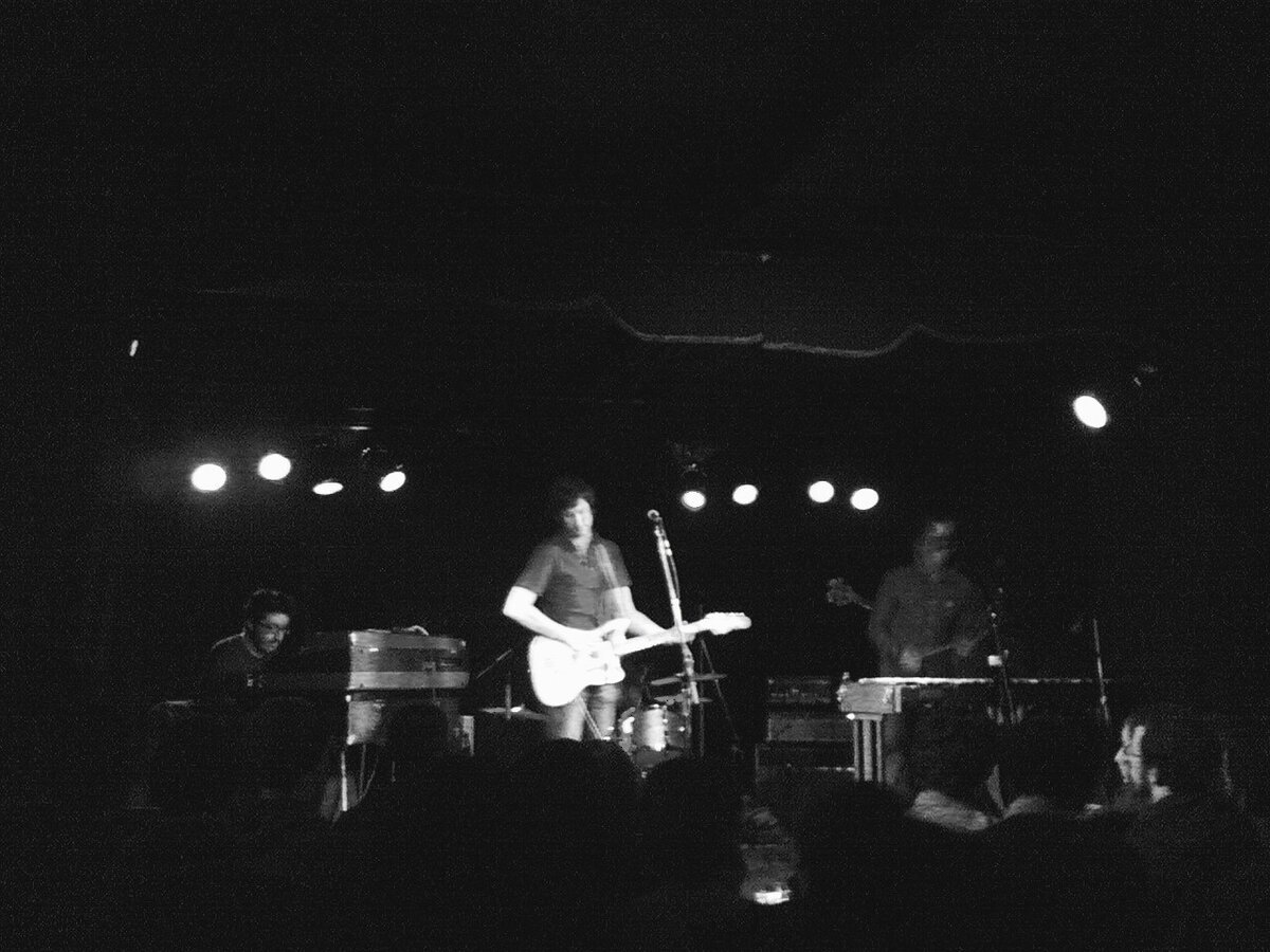 Black-and-white photo of a band playing on a dim club stage under small spotlights