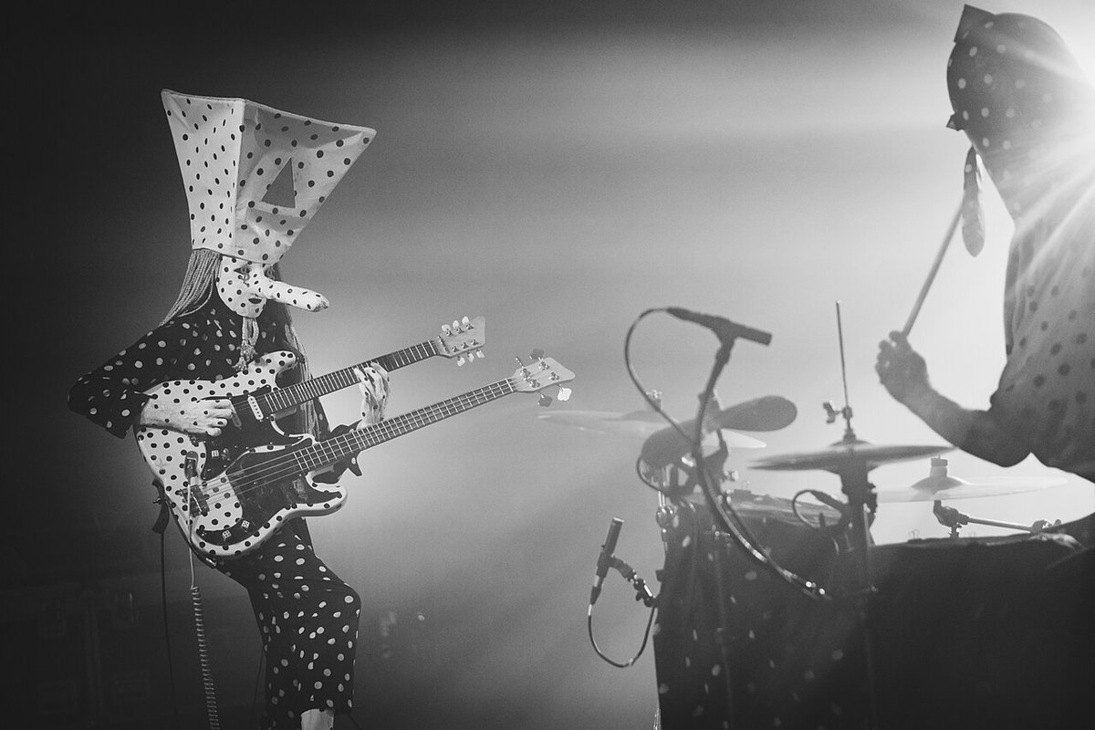 Black-and-white photo of Angine de Poitrine on stage, the guitarist in a polka-dot suit and tall pointed mask playing a double-neck guitar while the masked drummer plays in a burst of backlight