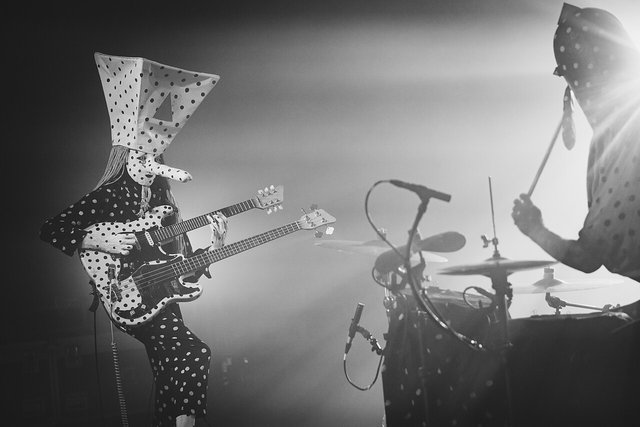 Black-and-white photo of Angine de Poitrine on stage, the guitarist in a polka-dot suit and tall pointed mask playing a double-neck guitar while the masked drummer plays in a burst of backlight