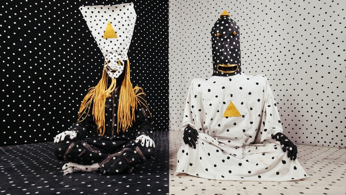 The two members of Angine de Poitrine seated in black-and-white polka-dot costumes and tall masks with gold triangles, one against a black dotted backdrop and one against a white one