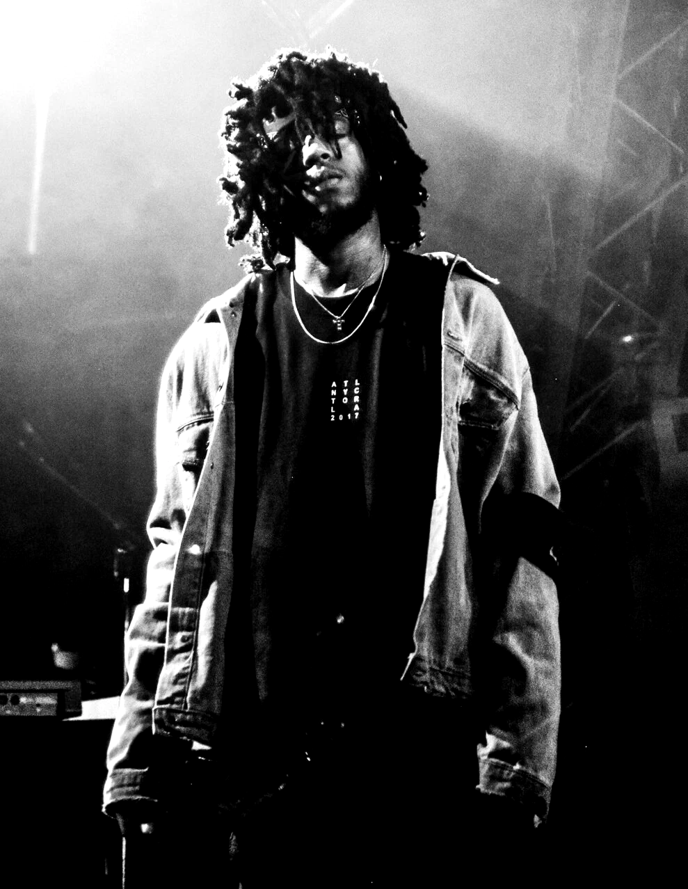 Black and white photo of 6LACK on stage in a denim jacket and chain necklace, hair falling over his face